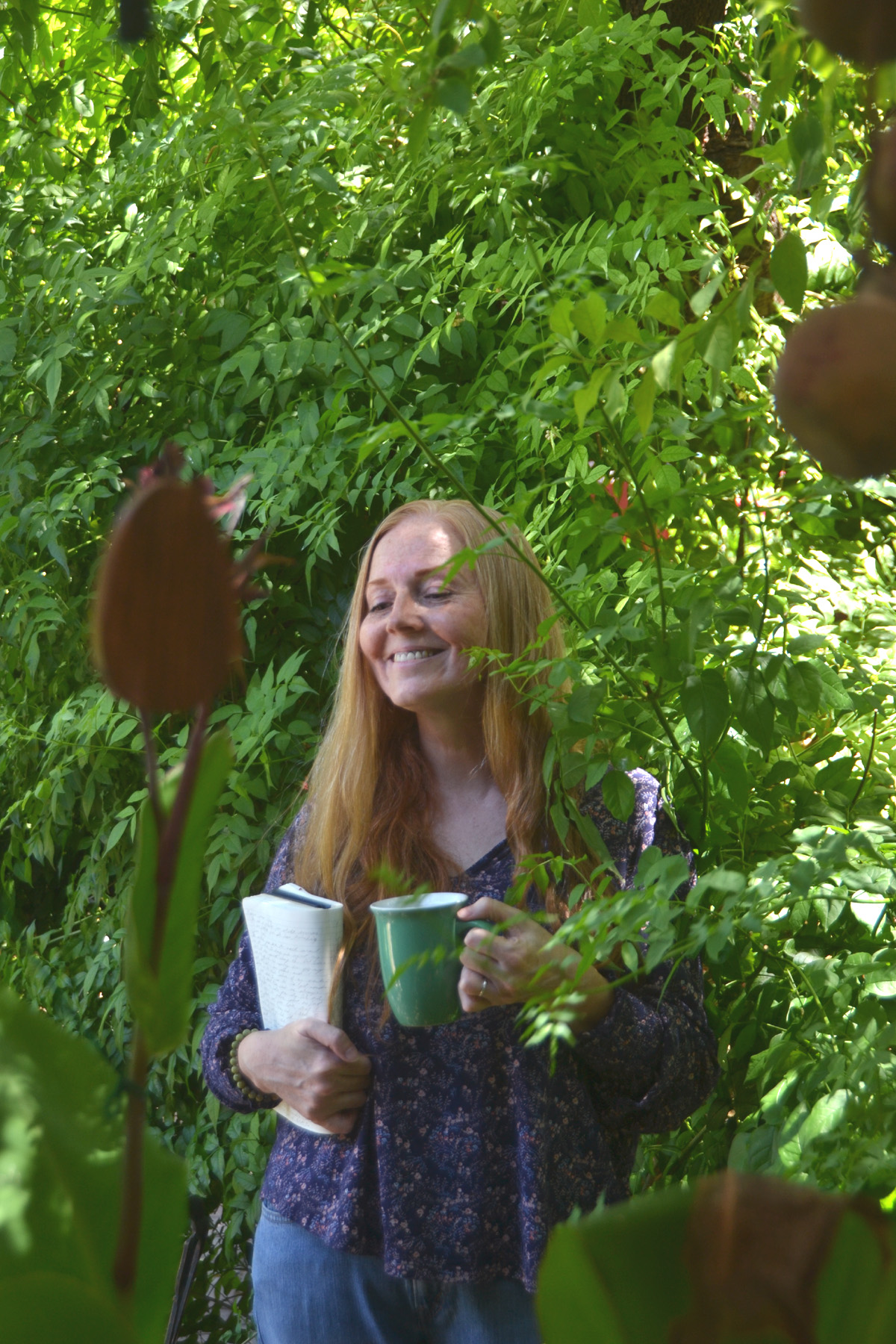 Dee smiling in the garden holding a cup of coffee and a journal.