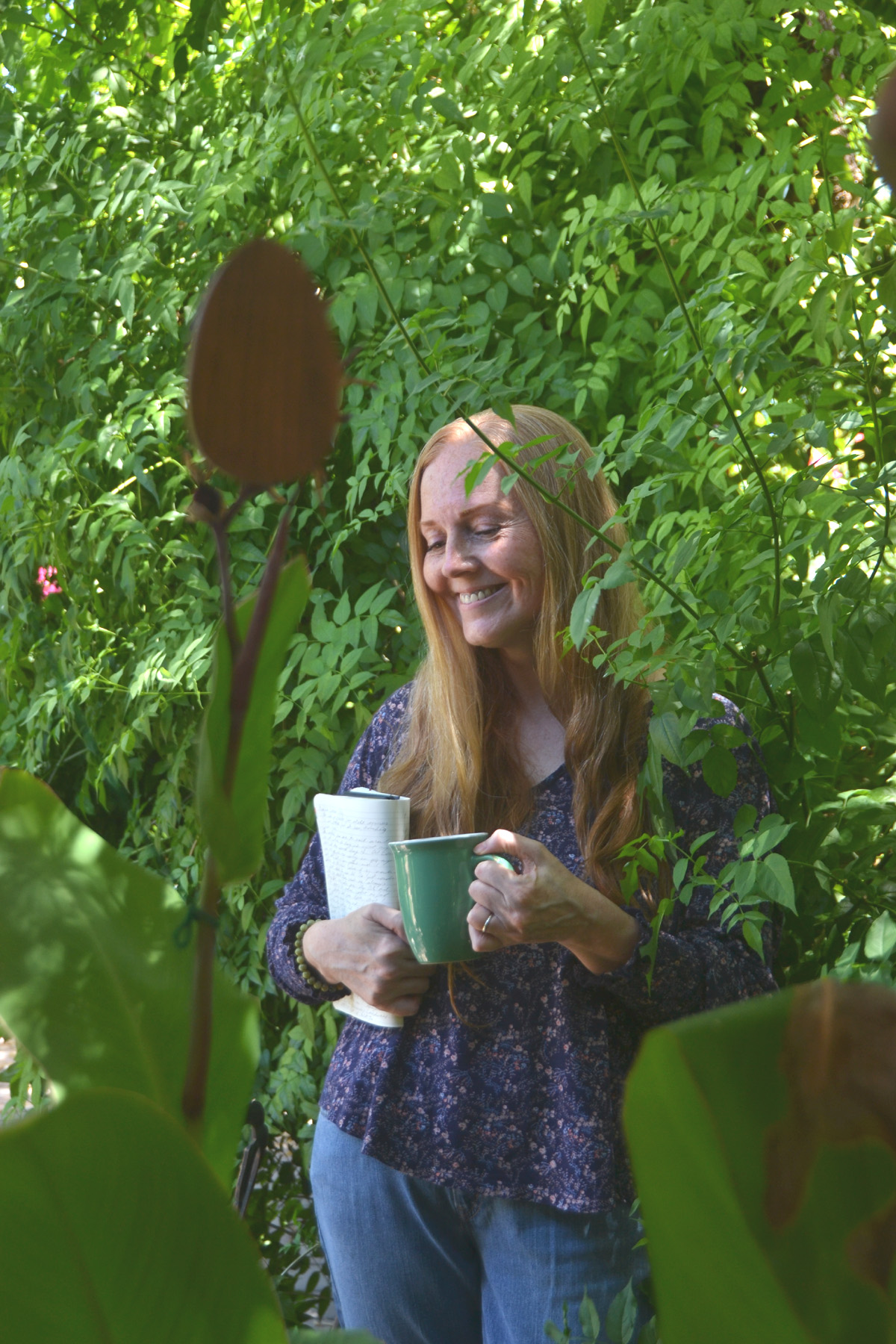 Dee stands near a tree holding a coffee cup and a journal.