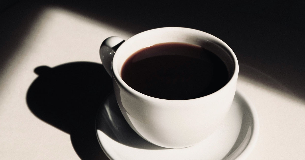 A cup of coffee stands on a sunlit white table in an example of how to become a morning person.