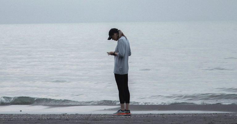 A solitary woman stands on the beach looking down at her phone in an example of tech addiction.