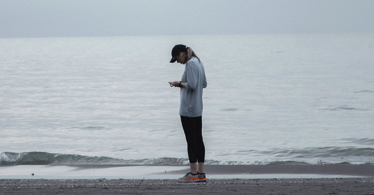 A solitary woman stands on the beach looking down at her phone in an example of tech addiction.