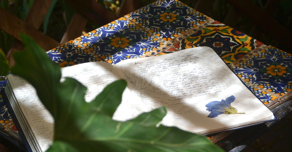 A notebook lays open on a table in a garden surrounded by greenery.